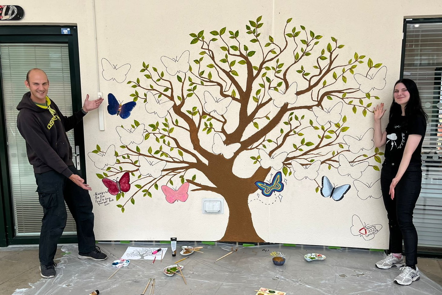 Adam and Carrie presenting their artwork. The mural is a brown tree trunk with green leaves and butterflies surrounding it