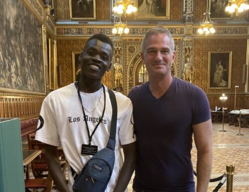 Nader stood next to Peter Kyle MP, both smiling wearing tshirts. They are stood in the houses of parliament in a grand room with golden walls. 