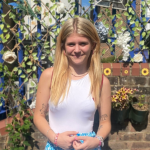 Alysha, a young person we supported. She is stood in the garden smiling against a flowery wall