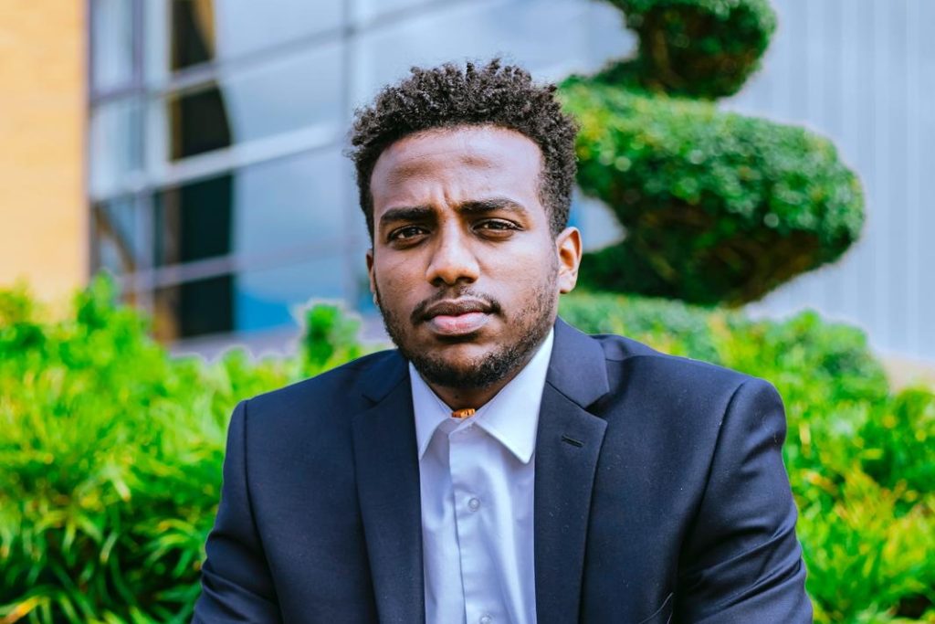 Amanuel, a young man wearing a suit and white shirt with green bushes behind him