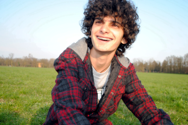 Dylan with curley brown hair and braces smiling at the camera, sat in a grass field